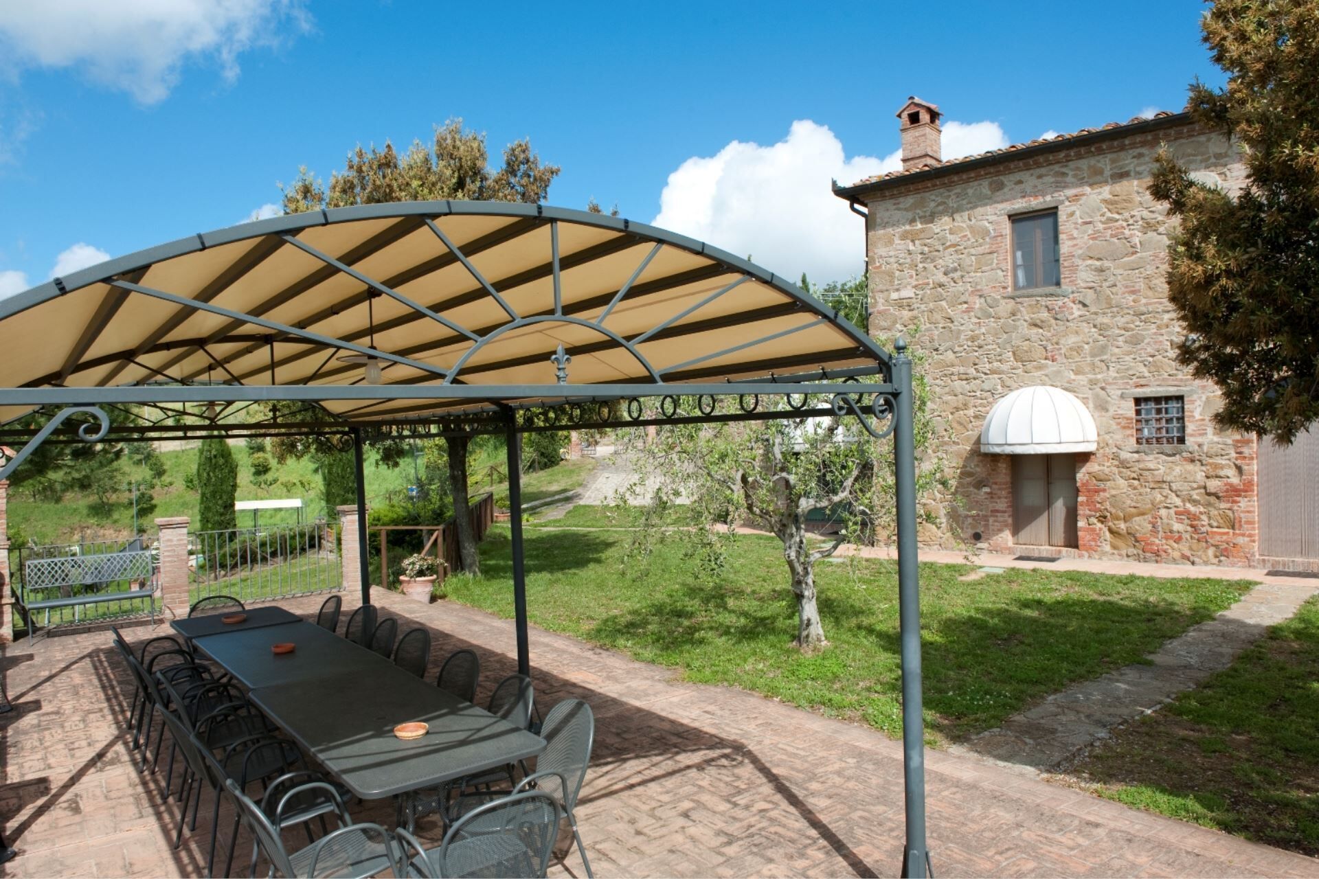 A covered outdoor seating area with a table and chairs, a stone building with a dome window, and a clear blue sky.