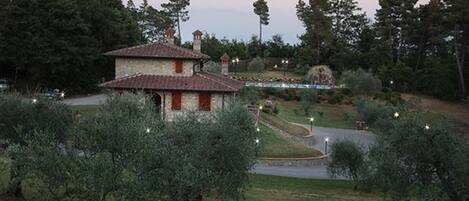 A rustic stone building with a tiled roof, surrounded by olive trees and a landscaped garden with a pathway.