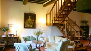 A cozy living room with a wooden staircase, a painting on the wall, and a small round table with a vase of flowers. - Olivo (Arezzo)