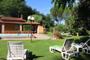 Un espace aménagé au bord de la piscine avec des chaises longues, un petit bâtiment doté d’un toit recouvert de tuiles rouges et un jardin où poussent différentes plantes et fleurs.