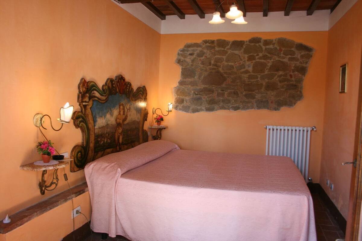 A bedroom with a stone wall, a bed with a pink bedspread, a nightstand with a lamp, and a small table with a flower vase.