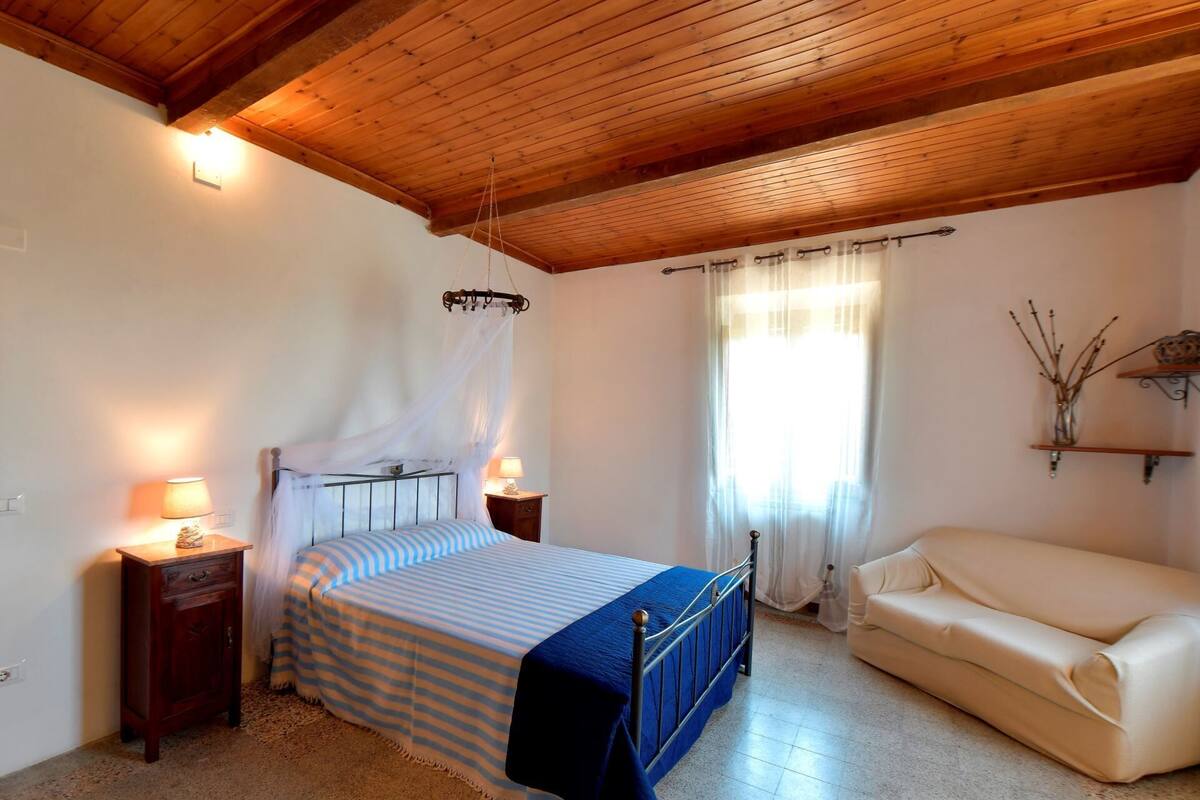 A bedroom with a bed, bedside tables, a nightstand, a window with curtains, a wooden ceiling, and a beige sofa.