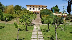 A large, two-story building with a tiled roof, surrounded by a well-maintained garden and a stone pathway leading to the entrance. - Poggio Falcone (Castiglione del Lago)