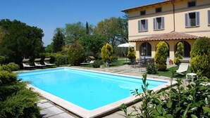 A swimming pool surrounded by lounge chairs and umbrellas, with a building featuring arched windows and a tiled roof in the background. - Poggio Falcone (Castiglione del Lago)