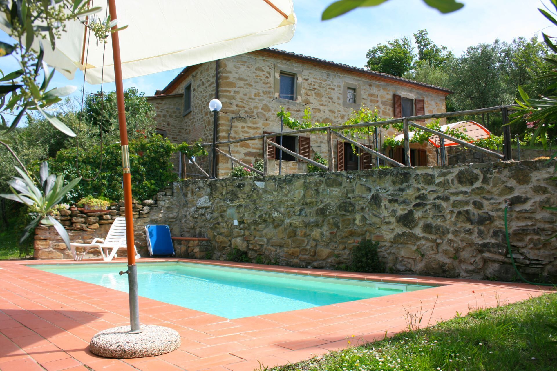 A stone house with a swimming pool, a white umbrella, and a blue chair.