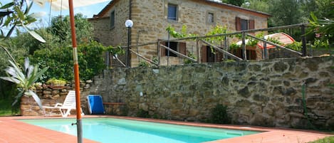 A stone house with a swimming pool, a white umbrella, and a blue chair.