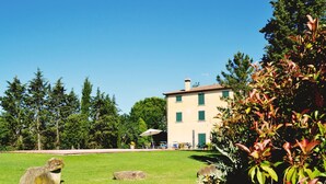 A building with a light-colored facade, multiple windows with green shutters, and a chimney. There are several large rocks in the foreground and a grassy area with trees in the background. - Villa Cantagallo (Cortona)
