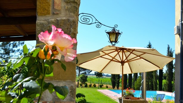 A stone pillar with a hanging lantern and a large beige umbrella in a garden setting. - Villa Cantagallo (Cortona)