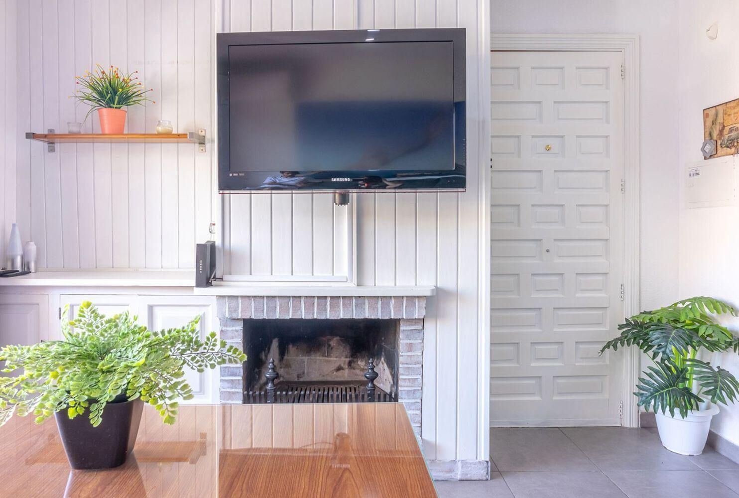 Ein Wohnzimmer mit Flachbildfernseher über einem Kamin, einem Holztisch und zwei Topfpflanzen.