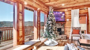 Un acogedor interior de cabaña con un gran árbol de Navidad, pisos de madera, una chimenea de piedra y un balcón con vistas panorámicas.