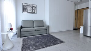 A modern living room with a grey sofa, a small table with a lamp, a framed picture on the wall, and a kitchen area with a refrigerator. - Victus Apartamenty - Cadena 3 (Sopot)