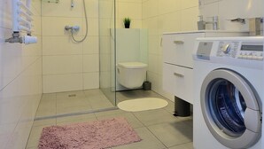 A modern bathroom with a glass shower enclosure, a white toilet, a washing machine, and a pink rug on the floor. - Victus Apartamenty - Cadena 3 (Sopot)
