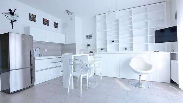 A modern kitchen with white cabinetry, a stainless steel refrigerator, and a dining area with clear chairs. - Victus Apartamenty - Cadena 3 (Sopot)