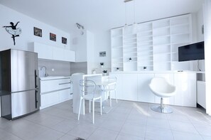 A modern kitchen with white cabinetry, a stainless steel refrigerator, and a dining area with clear chairs. - Victus Apartamenty - Cadena 3 (Sopot)