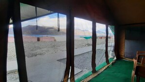 A room with a view of a mountainous landscape through large windows. There are two wooden chairs and a green carpet on the floor. - TIH P3 Camp - Pangong (Leh)