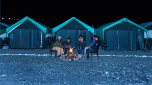 A group of people sitting around a campfire in front of a row of tents with a rocky ground. - TIH P3 Camp - Pangong (Leh)
