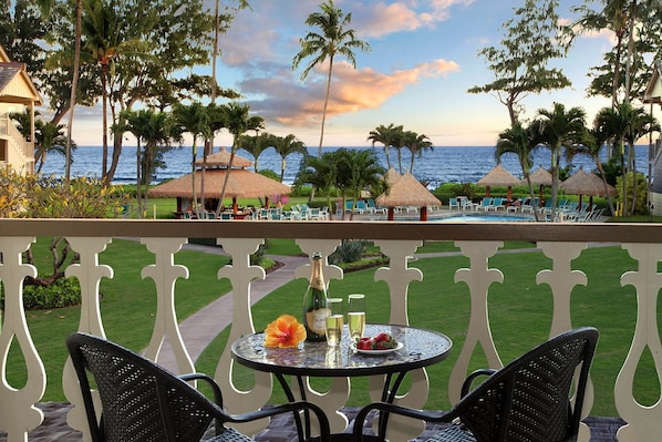 Un balcon avec une table dressée pour deux personnes, donnant sur un espace piscine agrémenté de chaises longues et de parasols, et offrant une vue sur l’océan.