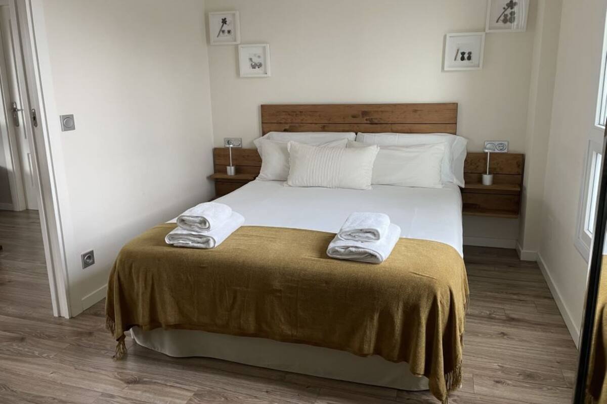 A neatly made bed with a brown bedspread and two folded towels, wooden headboard, white pillows, bedside tables with lamps, and framed pictures on the wall.