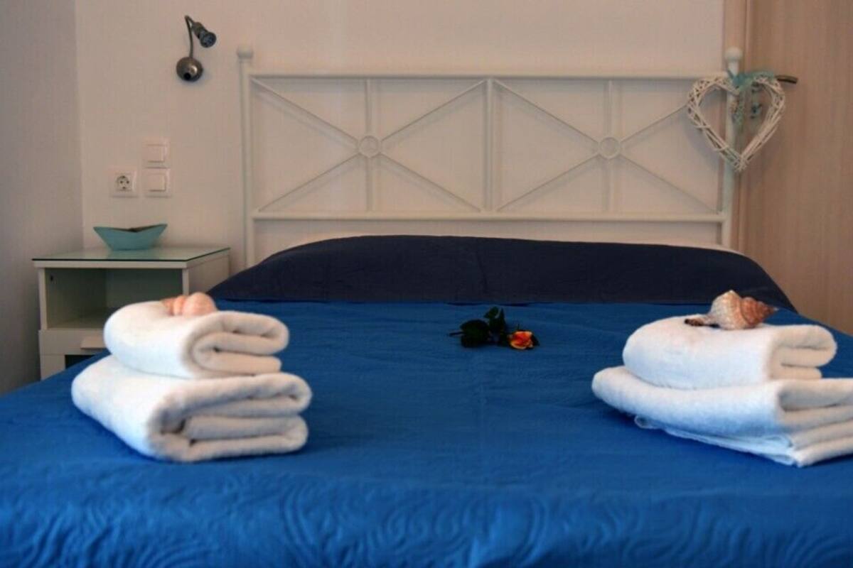 A neatly made bed with blue bedding, two folded white towels, a heart-shaped decoration, and a small plant on a bedside table.