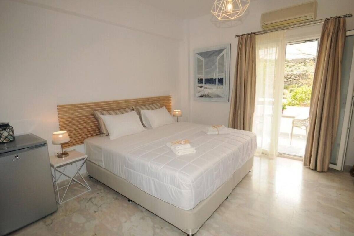 A bedroom with a bed, bedside tables, a small refrigerator, and a view of the outdoors.