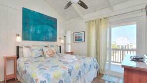 Une chambre à coucher avec un lit, un ventilateur de plafond, un grand tableau, une fenêtre avec des rideaux et une vue sur une maison à l’extérieur.