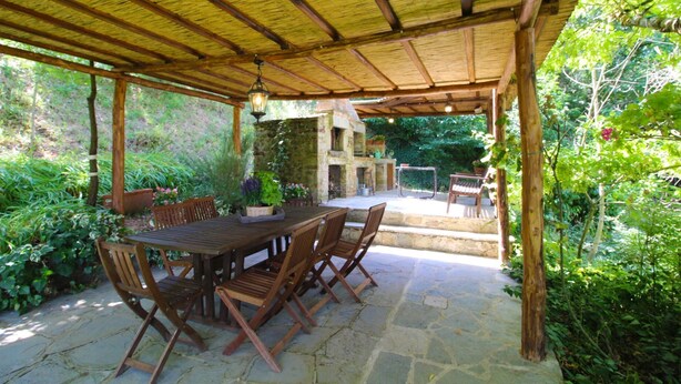 An outdoor patio with a stone floor, wooden furniture, and a stone fireplace.
