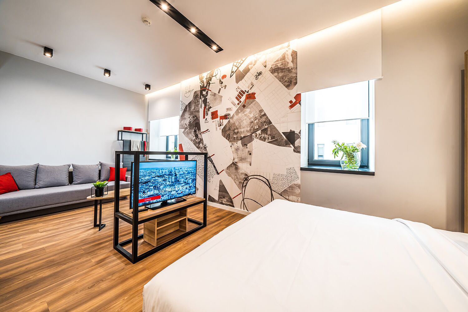 A modern hotel room with a large bed, a TV on a stand, a sofa, and a wall with a cityscape mural.
