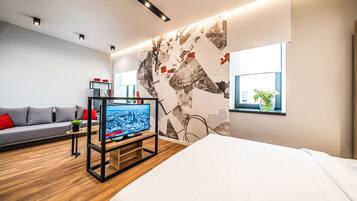 A modern hotel room with a large bed, a TV on a stand, a sofa, and a wall with a cityscape mural.