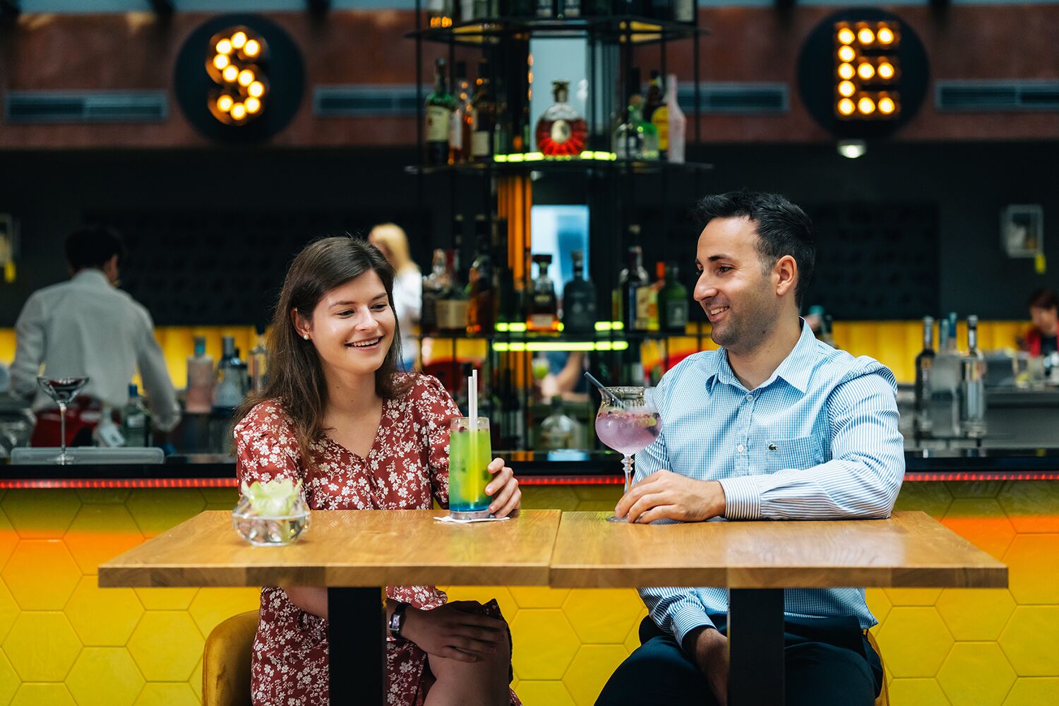 Two people seated at a table in a bar with a hexagonal floor pattern and a bar counter with various bottles in the background.