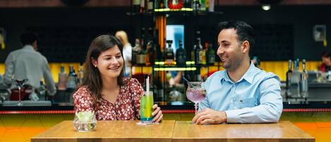 Two people seated at a table in a bar with a hexagonal floor pattern and a bar counter with various bottles in the background.