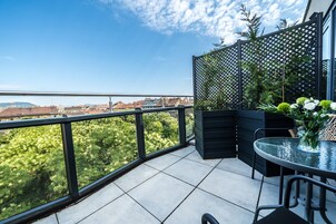 Un balcone con un tavolo e sedie in vetro, vista su vegetazione e edifici, e un pannello decorativo a griglia.