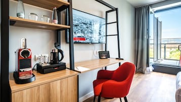 A modern hotel room with a wooden desk, a red chair, a coffee machine, and a large window offering a city view.
