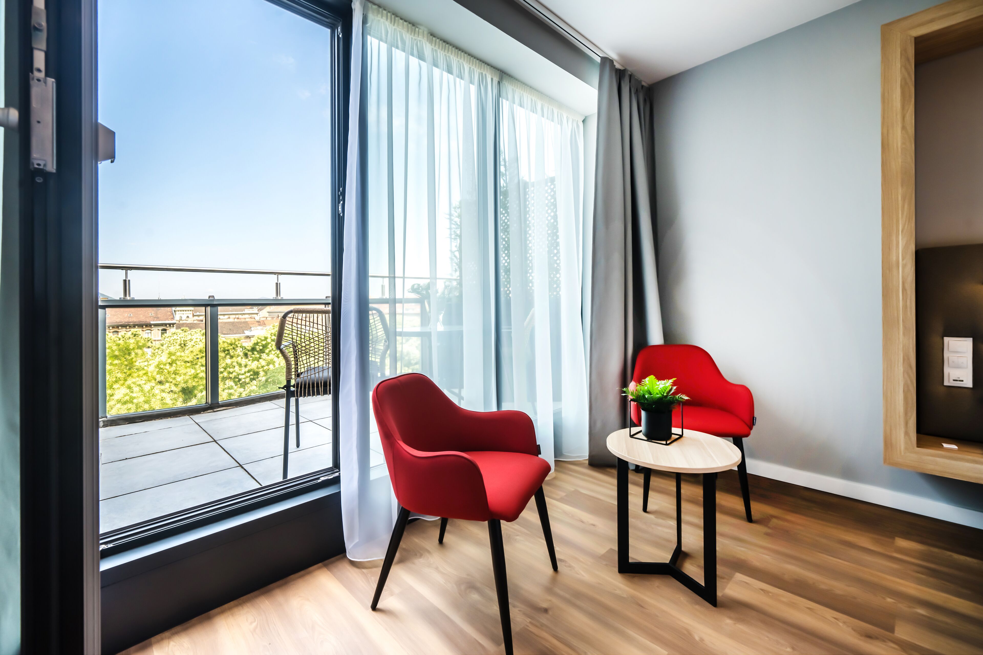 Une chambre moderne avec une grande fenêtre, deux fauteuils rouges, une petite table avec une plante et un plancher en bois.