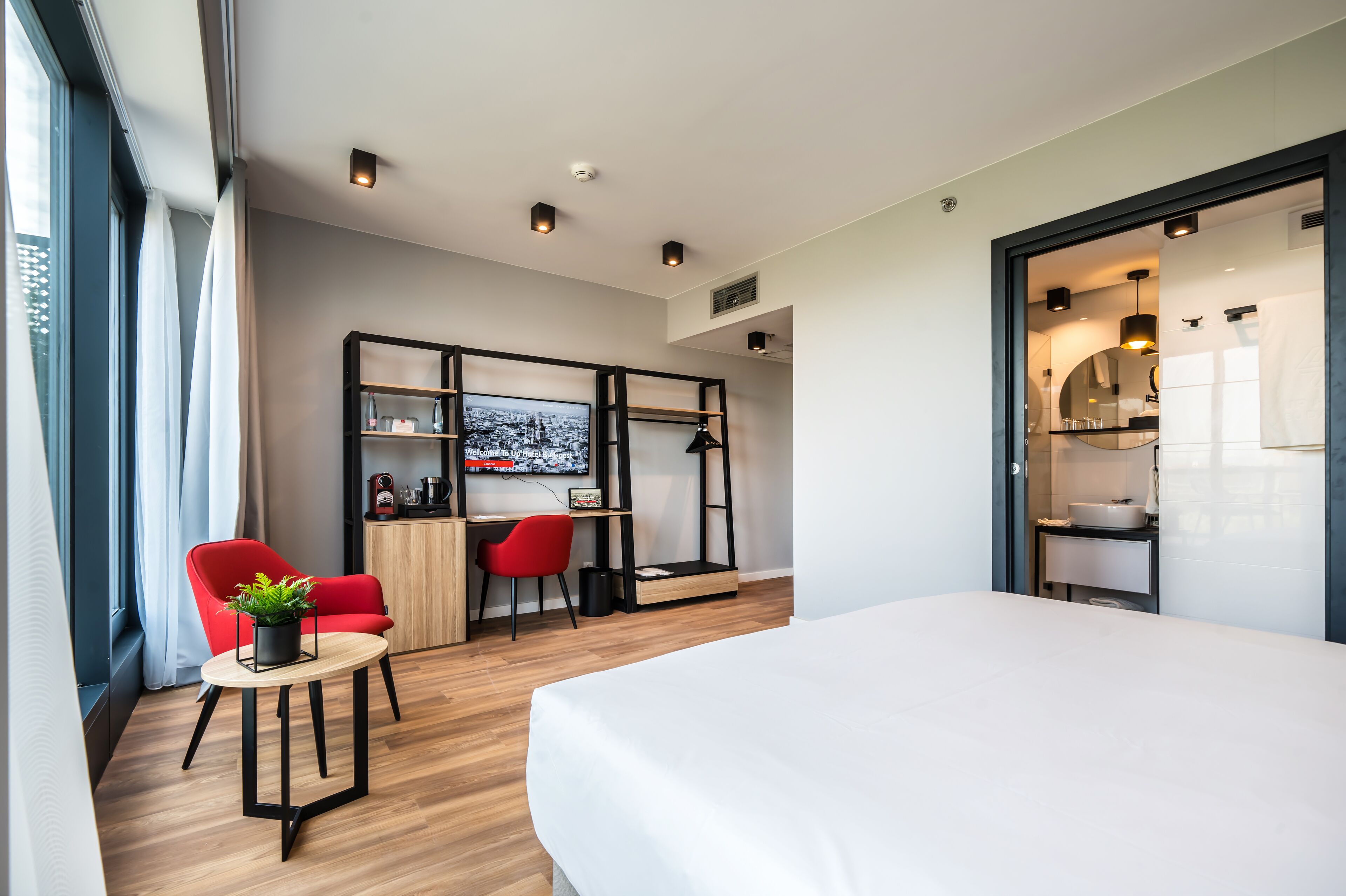 A modern hotel room with a large bed, a desk with a computer, a red chair, and a small table with a plant.