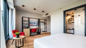 A modern hotel room with a large bed, a desk with a computer, a red chair, and a small table with a plant.