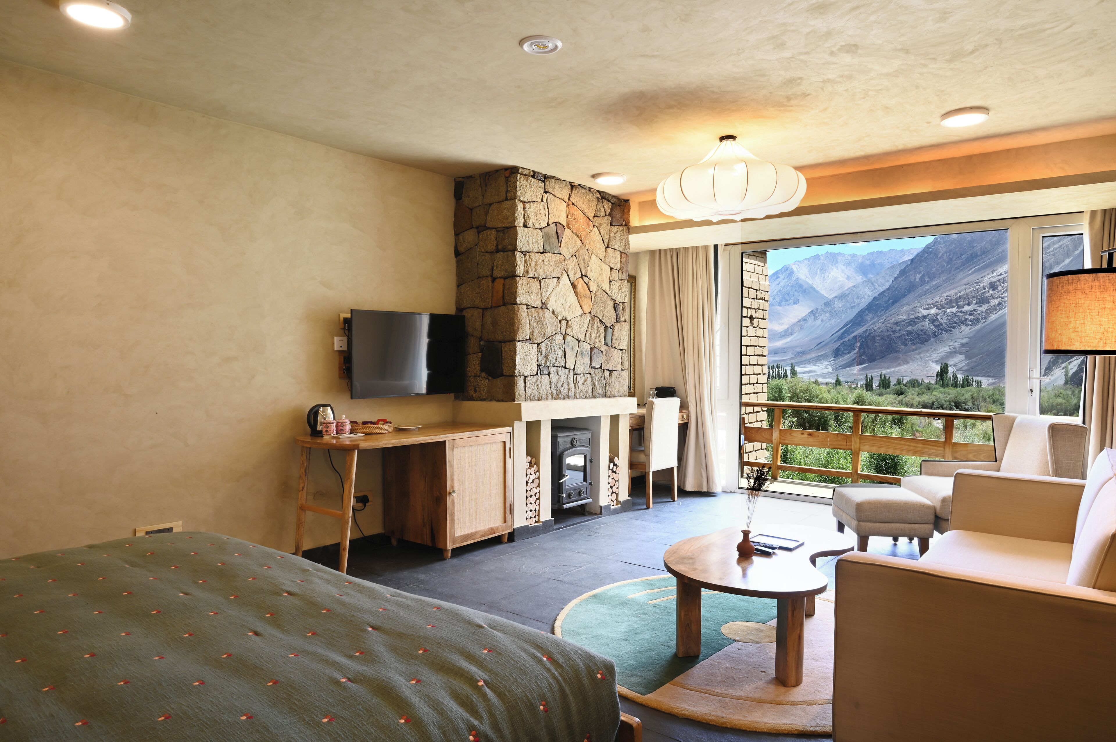 A hotel room with a stone fireplace, a television, a balcony with a mountain view, and a cozy seating area.