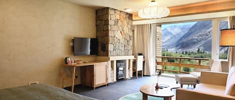 Habitación de hotel con chimenea de piedra, televisor, balcón con vista a la montaña y un rincón de estar acogedor.