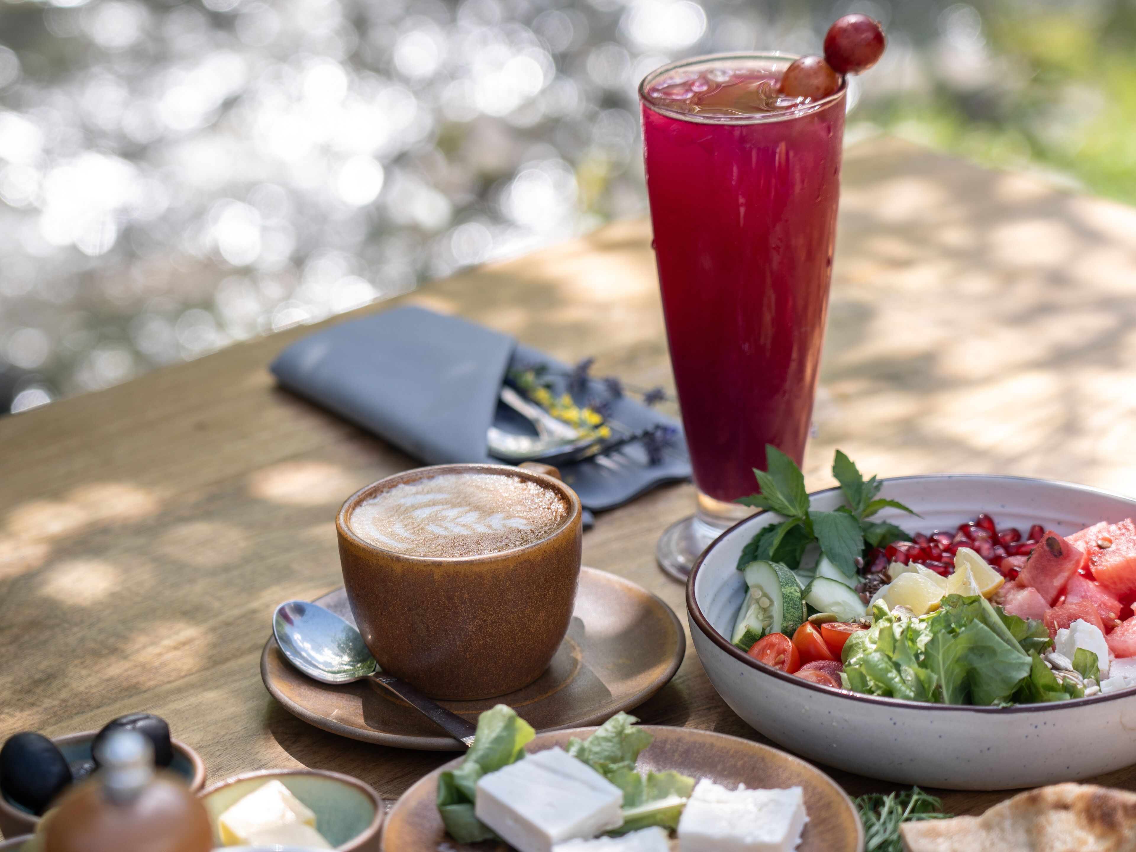 A table with a meal consisting of a bowl of salad, a cup of coffee, and a tall glass of red drink, with a napkin and spoon on the side.
