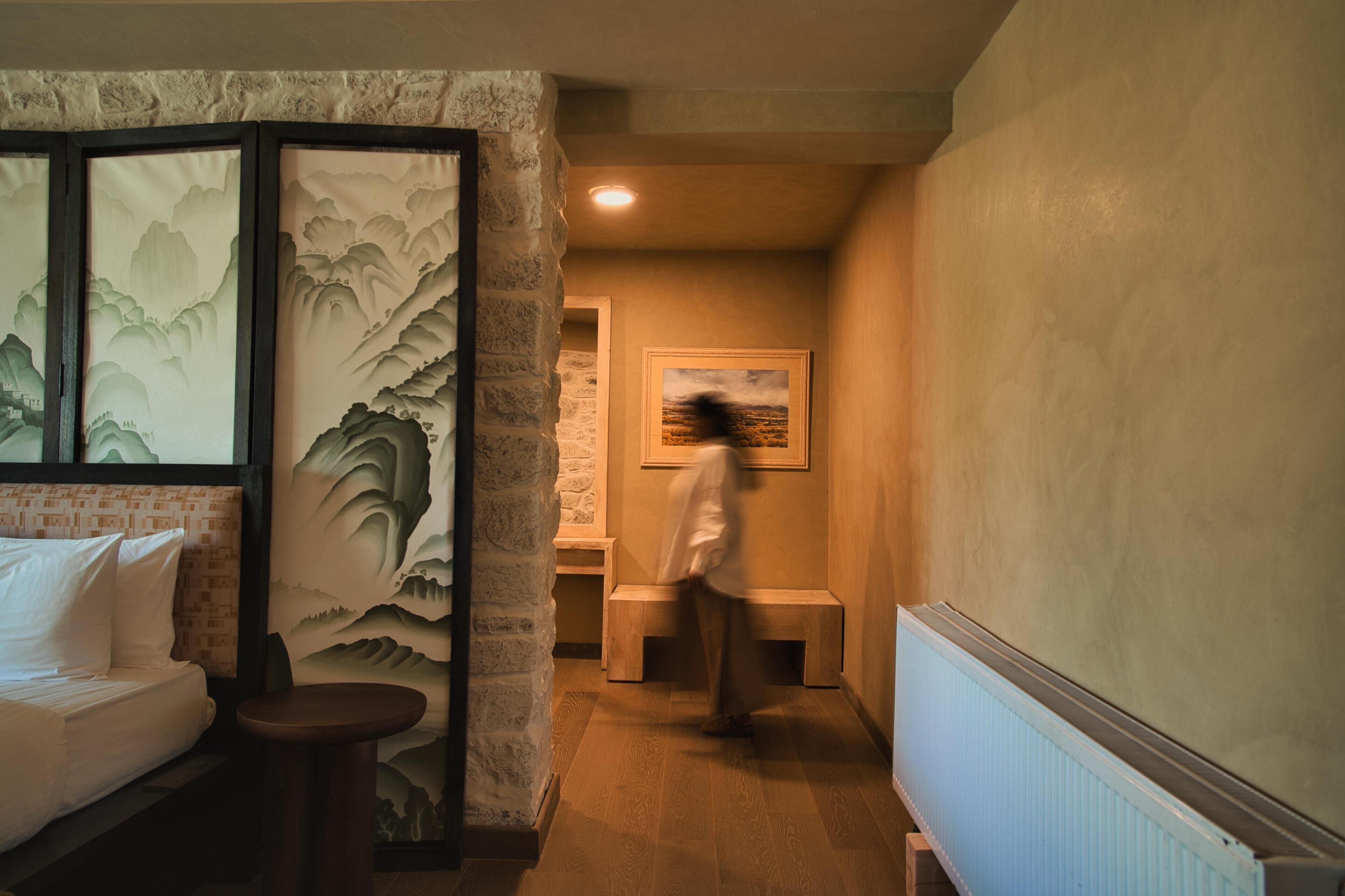 Une chambre d’hôtel avec un lit, un tabouret en bois, un paravent chinois traditionnel et un tableau accroché au mur.