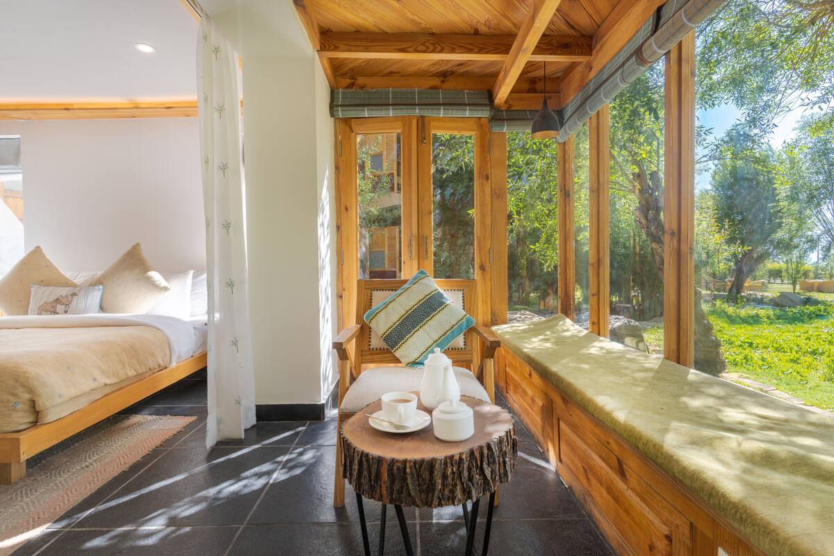 A wooden-framed room with a bed, a small table with a cup and a pitcher, and a view of greenery outside.