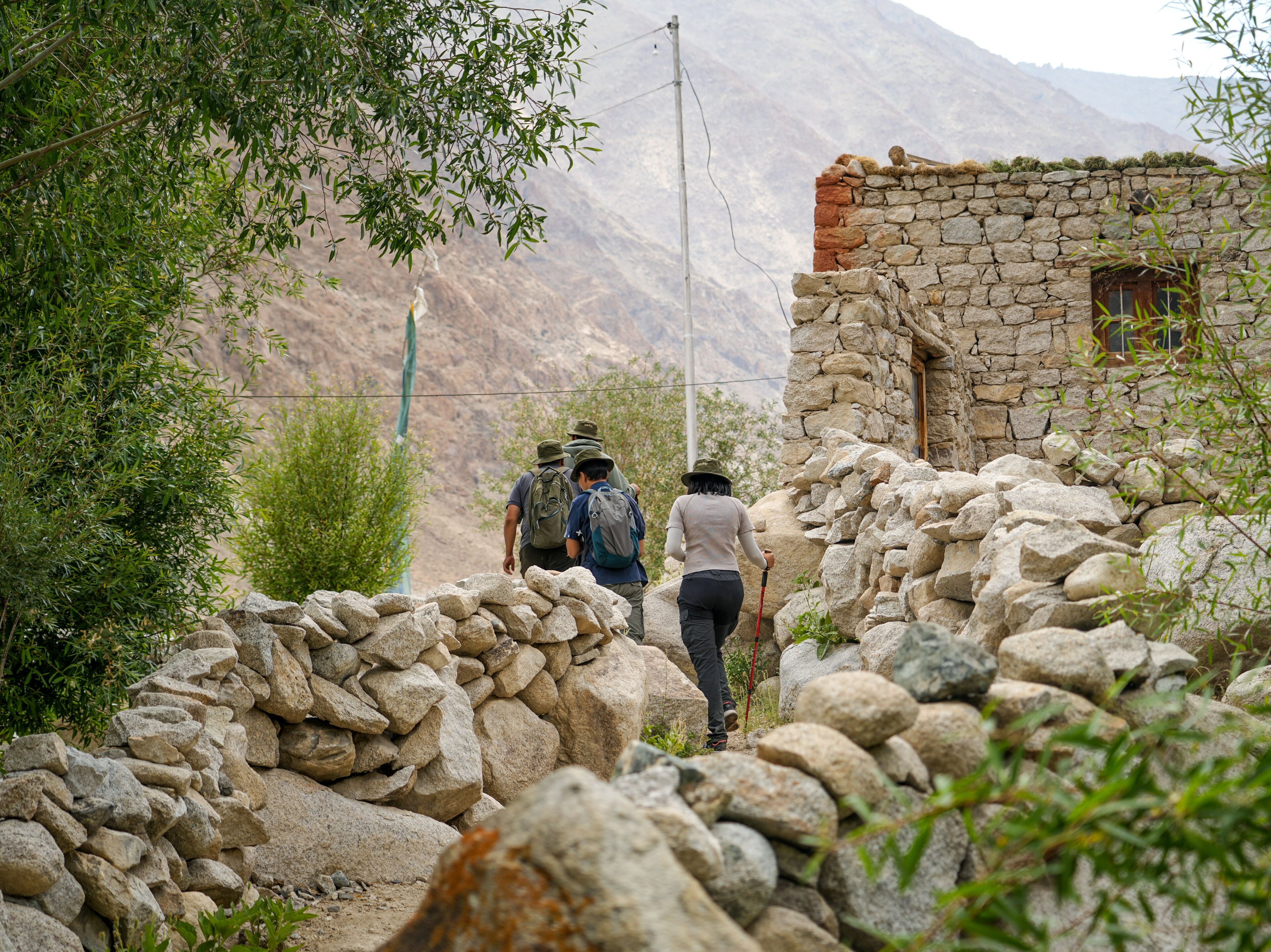 A narrow stone pathway leading to a rustic stone building with a wooden door, surrounded by greenery and mountains in the background.