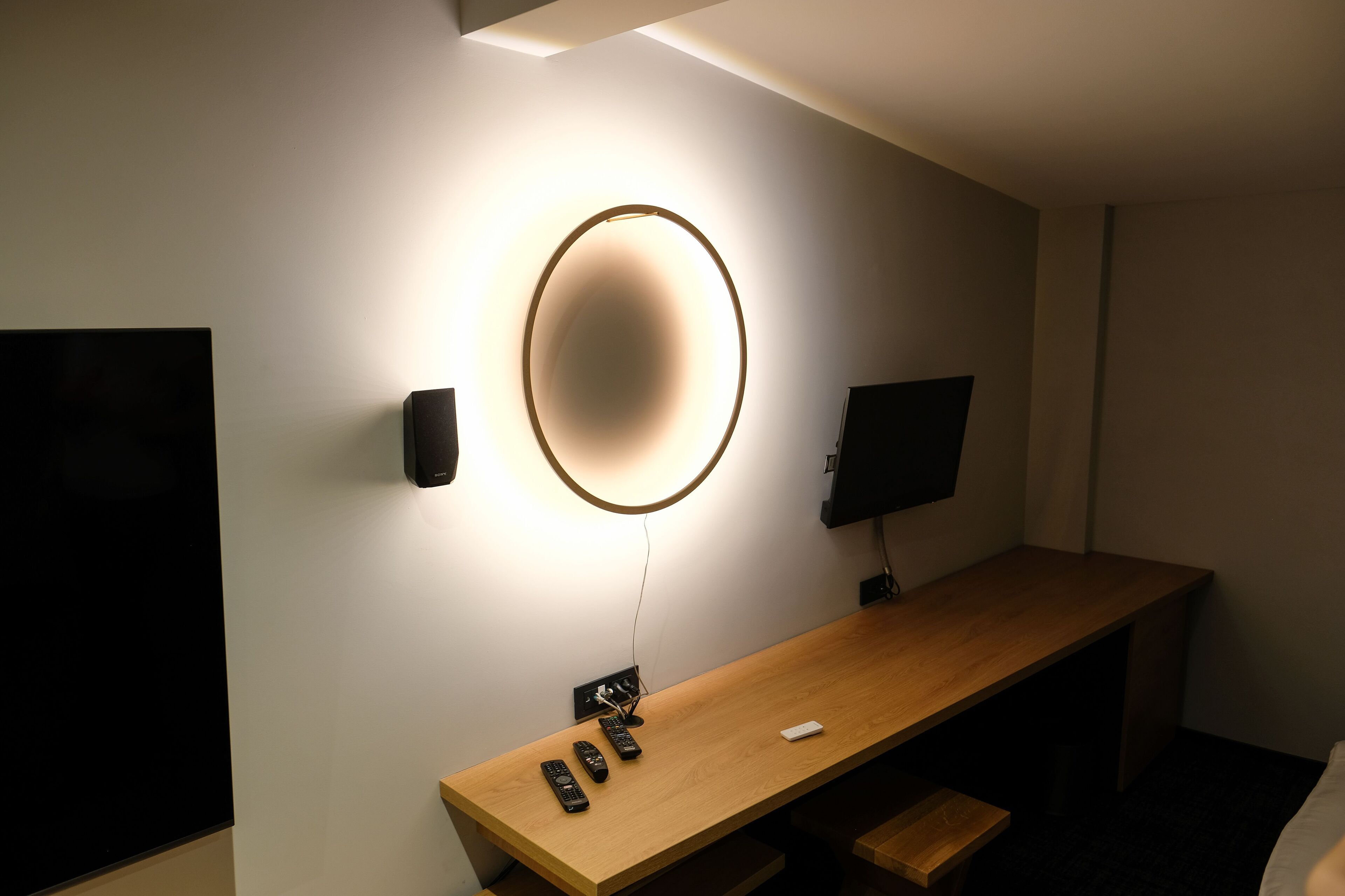 Habitació d'hotel amb escriptori de fusta, televisió, altaveu fixat a la paret i lluminària circular.