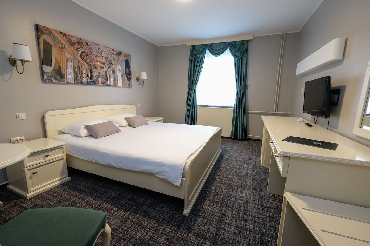 A hotel room with a large bed, a desk with a flat-screen TV, a painting on the wall, and a window with curtains.