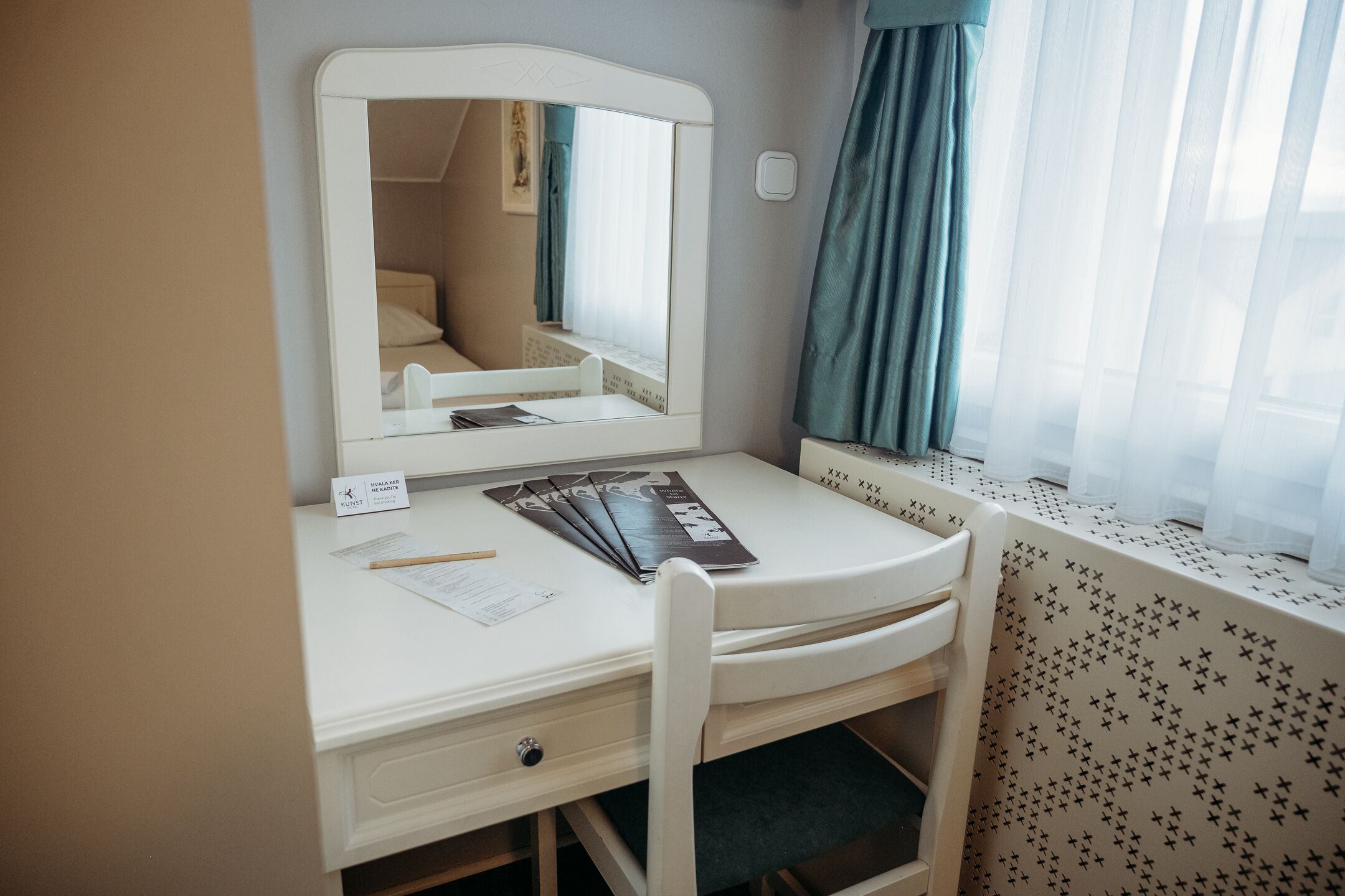 A small, neatly organized bedroom with a single bed, a bedside table, a mirror, and a window with sheer curtains.