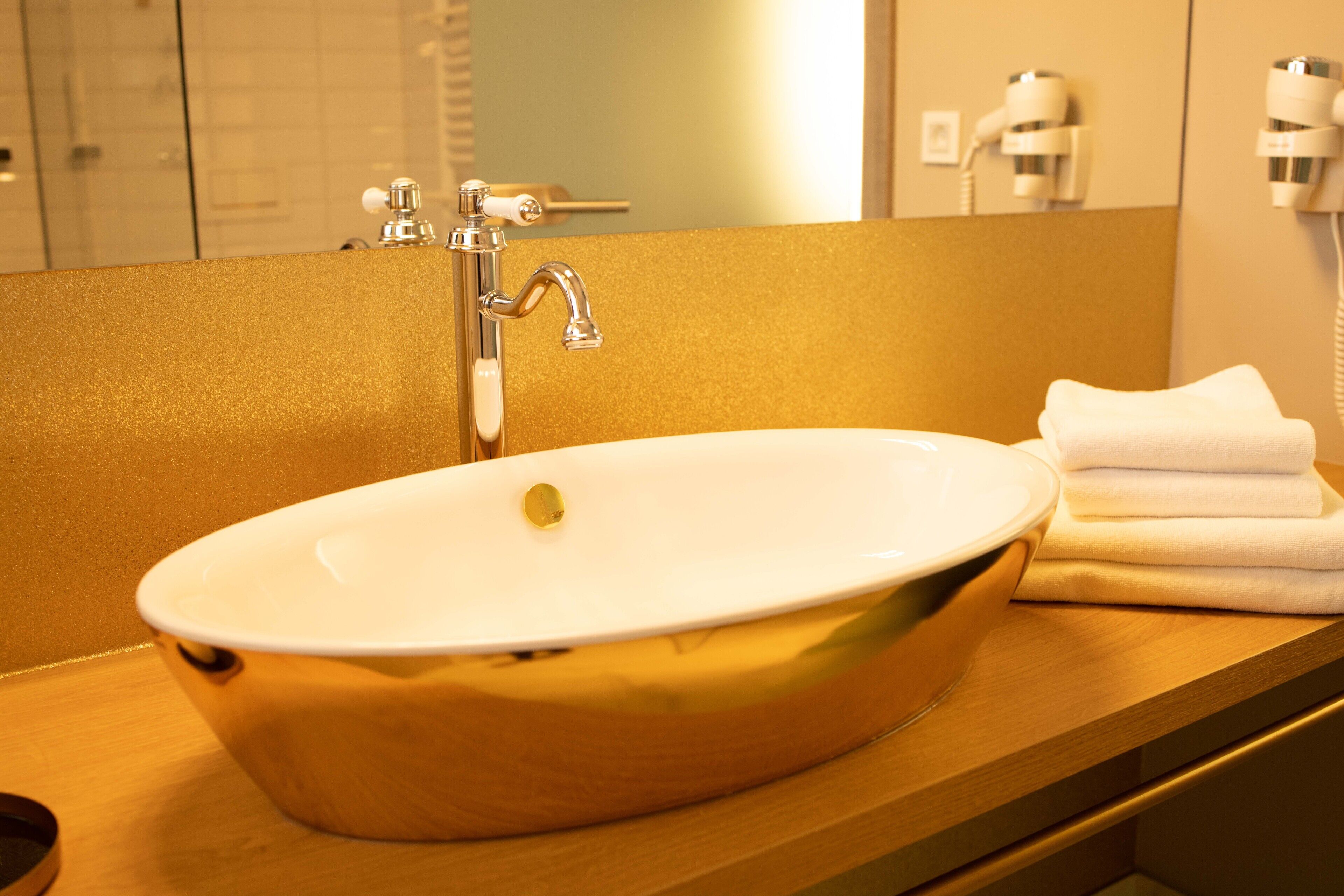 A bathroom with a white oval sink, a gold-colored countertop, and a mirror reflecting a hairdryer and a towel.