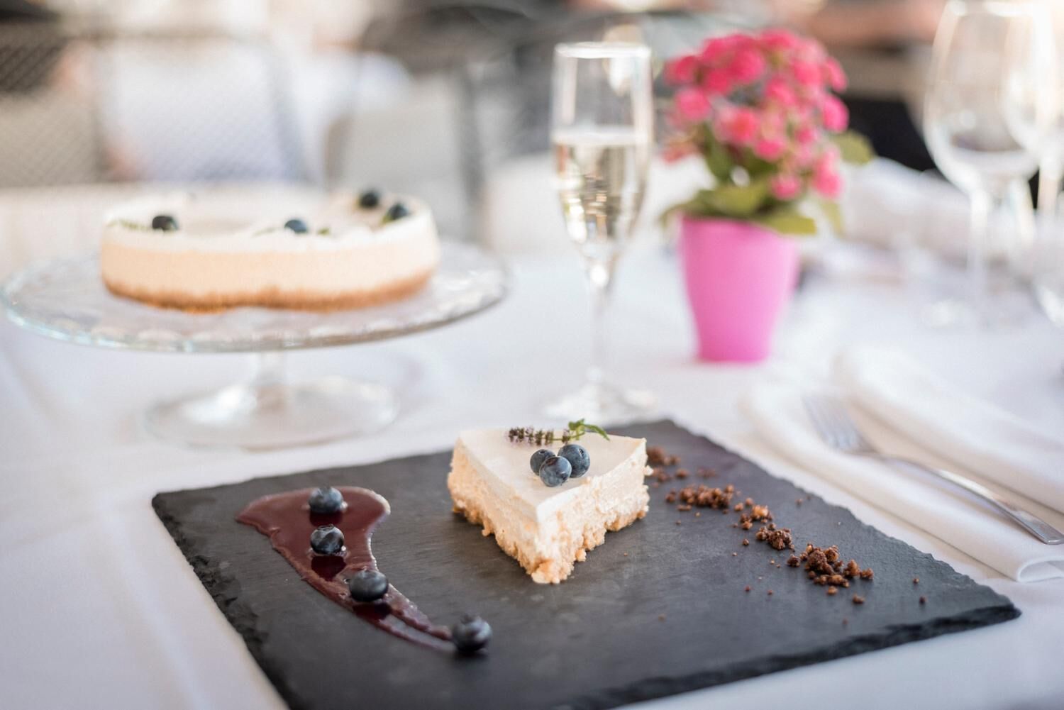Una mesa de postres con un pastel de queso y un postre de chocolate sobre un plato de pizarra, rodeada de copas de vino y un florero con una flor rosa.