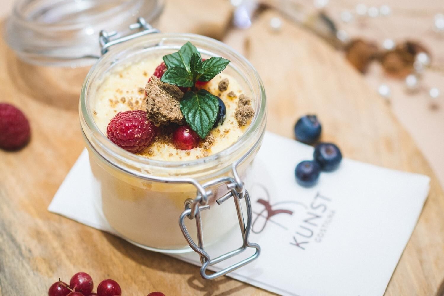 Postre en un frasco de vidrio con cierre metálico, adornado con una hoja de menta, frambuesas y arándanos, sobre una superficie de madera con una servilleta y un logotipo.