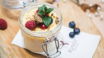 Postre en un frasco de vidrio con cierre metálico, adornado con una hoja de menta, frambuesas y arándanos, sobre una superficie de madera con una servilleta y un logotipo.