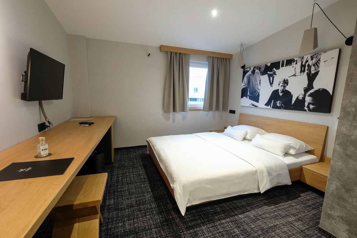 A hotel room with a bed, a desk, a TV, a window with curtains, and a large photo on the wall.