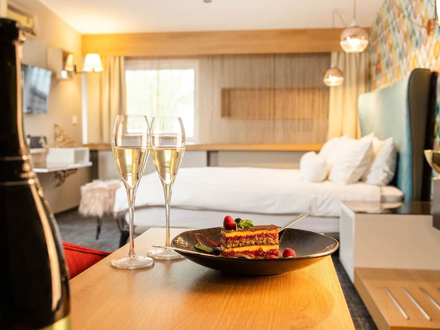 A dining table with a plate of cake and two glasses of wine, a bottle of wine, and a bed in the background.